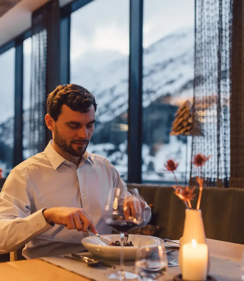 Gourmetrestaurant im hinteren Ötztal Mann sitzt an einem gedeckten Tisch im Hotelrestaurant und isst ein Gericht, auf dem Tisch Kerze, Weingläser und Vase, große Fenster mit Bergblick dahinter.