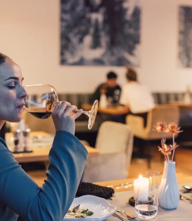 Gourmetrestaurant im hinteren Ötztal Frau sitzt an einem gedeckten Tisch im Restaurant und trinkt aus einem Rotweinglas, im Hintergrund weitere Tische und Gäste in einem modern gestalteten Gastraum.